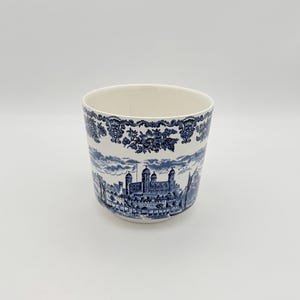 May include: A white ceramic planter with a blue and white transferware design. The design features a detailed illustration of a castle and floral patterns. The planter has a cylindrical shape and a smooth, glossy finish.