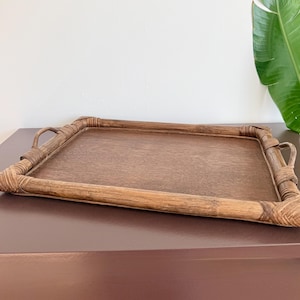 Peut inclure: Un plateau de service rectangulaire fabriqué en rotin tressé marron. Le plateau présente une base plate et lisse ainsi qu'un rebord surélevé avec des coins renforcés. Deux petites poignées arquées sont fixées sur les côtés courts.