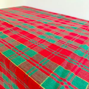 May include: A rectangular fabric tablecloth featuring a vibrant plaid pattern with intersecting lines of bright red, forest green, and thin gold metallic threads woven throughout the textile to create a repeating checkered design.