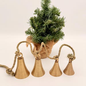 May include: Four gold-colored bells with a textured surface are strung together with a rope. A small artificial Christmas tree in a burlap pot is in the background. The bells are a decorative item.
