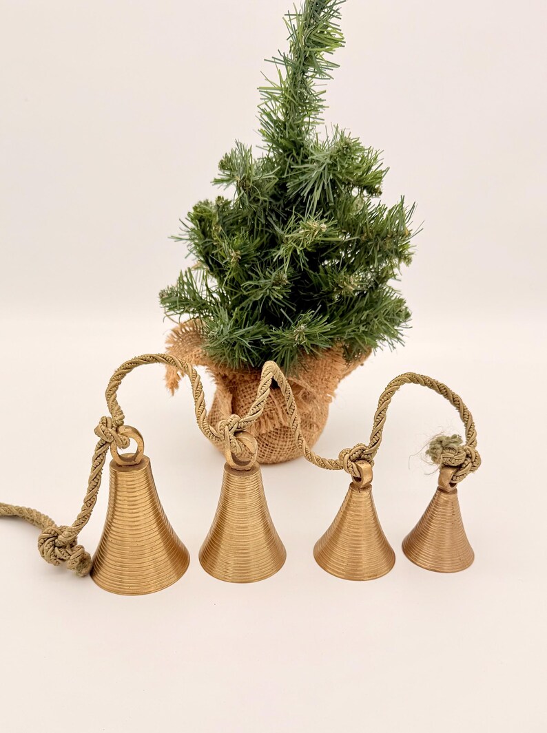 May include: Four gold-colored bells with a textured surface are strung together with a rope. A small artificial Christmas tree in a burlap pot is in the background. The bells are a decorative item.