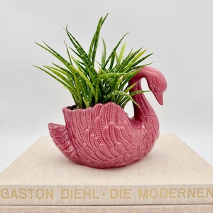 Op de afbeelding: Een glanzende, donkerroze keramische plantenbak in de vorm van een zwaan met gedetailleerde, gevormde veerstructuren op het lichaam. De zwaan bevat een levendige groene sansevieria met puntige, gevlekte bladeren die uit het midden omhoog steken.