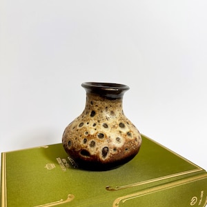 Könnte beinhalten: Eine kleine Keramikvase mit einem gesprenkelten Muster in Braun- und Beigetönen. Die Vase hat einen runden Körper und einen dunkelbraunen Rand. Sie steht auf einem grünen Buch.