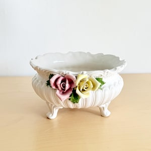 May include: White ceramic planter with a scalloped rim and three small feet. The planter is decorated with two ceramic roses, one pink and one yellow, with green leaves. A decorative piece for plants or flowers.