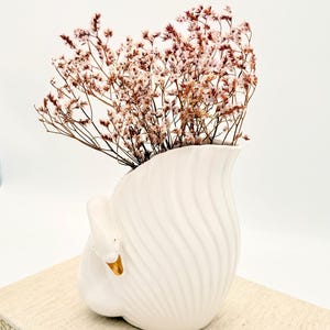 May include: A white ceramic swan-shaped vase with a gold beak and accents, holding dried pink flowers. The vase has a textured, swirled design. The vase is sitting on a light-coloured surface.