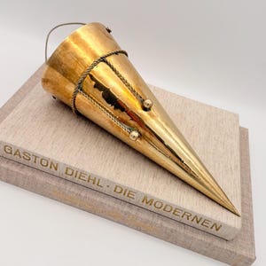 May include: A gold-colored, cone-shaped decorative object with a handle and rope detailing. The object is resting on top of two books with the text "GASTON DIEHL - DIE MODERNEN" printed on the spine. The object has a shiny, metallic finish.
