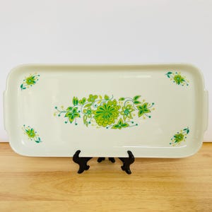 May include: An off-white rectangular serving tray with a floral pattern in green and yellow. The tray has rounded corners and small handles. The floral design is centered and repeated in each corner.