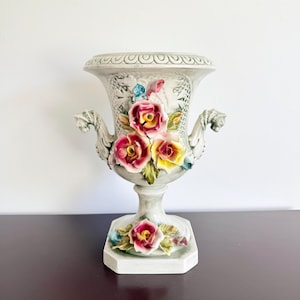 May include: A ceramic urn-style vase featuring a pale gray finish with a textured lattice pattern. The piece is adorned with three-dimensional hand-painted pink and yellow roses and green leaves, with two ornate side handles and a hexagonal base.