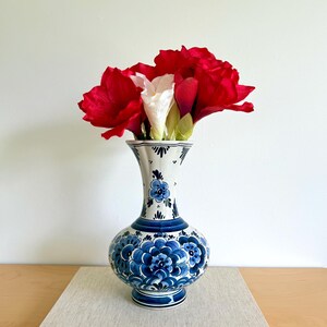 May include: A white and blue ceramic vase with a floral design, holding red and white flowers. The vase sits on a light-colored rectangular block, with the text "GASTON DIEHL · DIE MODERNEN" visible.