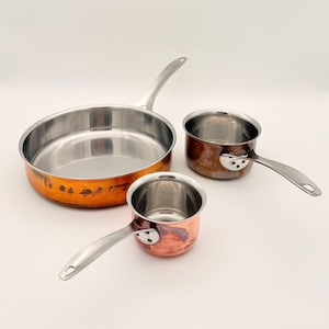 May include: Three copper-bottomed stainless steel cooking pots with silver handles. The largest is a frying pan, and the other two are saucepans. The copper has a warm, aged patina, and the interiors are shiny.