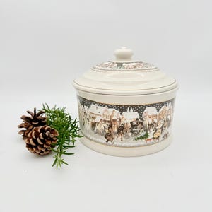 Op de afbeelding: Een ronde, crèmekleurige keramische pot met deksel, voorzien van een gedetailleerde illustratie van een besneeuwd dorpsgezicht met figuren, gebouwen en bomen. Twee bruine dennenappels en een takje groene rozemarijn liggen naast de pot.