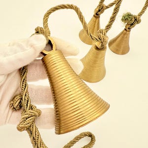 May include: A collection of brass bells with a textured, ribbed design. The bells have a conical shape and are suspended by twisted rope handles. The bells are a warm gold color and are displayed against a white background.
