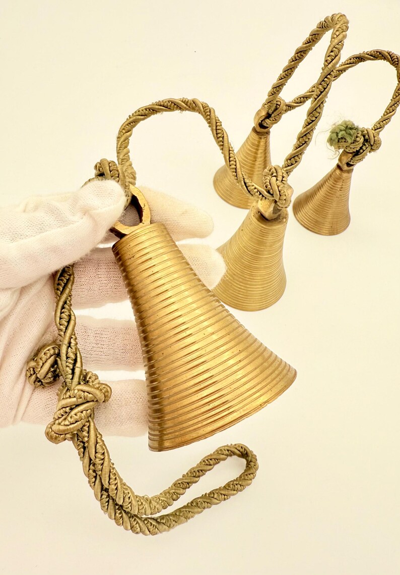May include: A collection of brass bells with a textured, ribbed design. The bells have a conical shape and are suspended by twisted rope handles. The bells are a warm gold color and are displayed against a white background.