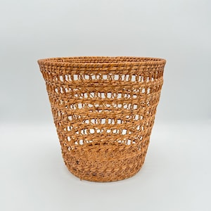Puede incluir: Una cesta tejida de forma cilíndrica. La cesta está hecha de mimbre marrón claro y tiene un tejido abierto, similar a una celosía. La cesta está vacía y se encuentra sobre un fondo blanco.