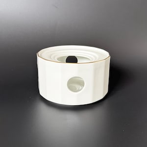 May include: A white ceramic tea warmer with a gold rim. The warmer has a geometric design with a circular opening on the side and a larger opening on top. The tea warmer is designed to hold a tea pot.