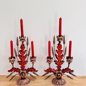 Könnte beinhalten: Paar verzierter rot-goldener Kerzenhalter mit roten Kerzen. Jeder Halter hat ein Blumen- und Blattdesign mit drei Kerzenhaltern und weizenartigen Akzenten. Die Kerzenhalter stehen auf einer Holzoberfläche.