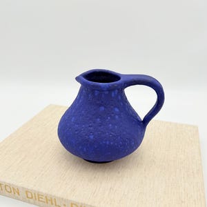 Puede incluir: Una pequeña jarra azul cobalto texturizada con asa y pico. La jarra de cerámica tiene un aspecto moteado y se encuentra sobre una superficie de color claro. La jarra es un objeto decorativo.