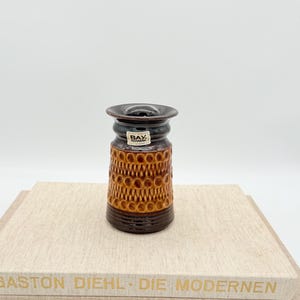 Puede incluir: Un pequeño jarrón de cerámica marrón con un diseño texturizado y estampado. El jarrón tiene una parte superior e inferior marrón oscuro, con un cuerpo marrón claro con un patrón de círculos repetidos. Una pequeña etiqueta está adherida al frente.