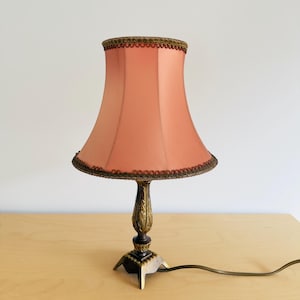 Pu&ograve; includere: Una lampada da tavolo vintage con paralume color pesca e base decorativa in bronzo. Il paralume &egrave; rifinito con un bordo decorativo marrone scuro. La lampada ha una base a tre gambe e un cavo di alimentazione. La lampada &egrave; su una superficie di legno chiaro.