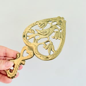 Può includere: Un sottopentola in ottone a forma di cuore caratterizzato da un design traforato con un uccello appollaiato tra fiori e foglie stilizzate. L'oggetto presenta un manico decorativo con un piccolo intaglio a forma di cuore.