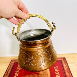 Op de afbeelding: Een koperkleurige metalen emmer met een sierlijk ontwerp en een messing handvat. De emmer heeft een zilveren binnenkant en wordt door een hand vastgehouden. De emmer staat op een rood boek.