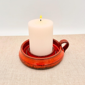 Puede incluir: Una vela de pilar blanca encendida se encuentra en un portavelas de cerámica roja. El portavelas tiene un diseño circular con un asa en el lateral. El portavelas es rojo con detalles marrones.