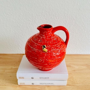 May include: A bright red ceramic jug with a textured finish and a handle. The jug features a small spout and a decorative detail. It is placed on two white books.