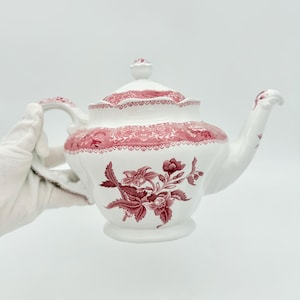Può includere: Una teiera in ceramica bianca con un motivo floreale rosso scuro. Il design presenta un'illustrazione botanica dettagliata sul corpo, un bordo decorativo rosso abbinato attorno al coperchio e al bordo superiore, un manico curvo e un beccuccio.