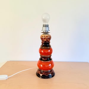 Könnte beinhalten: Eine Vintage-Tischlampe aus Keramik mit einer Glühbirne. Die Lampe hat ein gestapeltes Design mit glänzendem, rot-orangem Finish und dunkelbraunen Akzenten. Die Lampe ist eingesteckt und steht auf einer hellen Holzoberfläche.