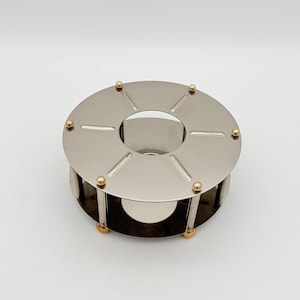 May include: A stainless steel fondue pot warmer with a circular design. The warmer has a round top with six cut-out sections and gold-colored accents. The base is also stainless steel, with gold-colored feet.