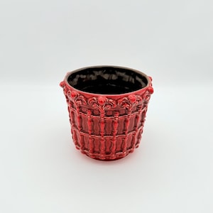 Puede incluir: Una maceta de cerámica roja con un diseño detallado y texturizado. La maceta tiene un interior oscuro y un borde decorativo. El exterior presenta un patrón repetido de elementos verticales y horizontales, creando un aspecto clásico y ornamentado.