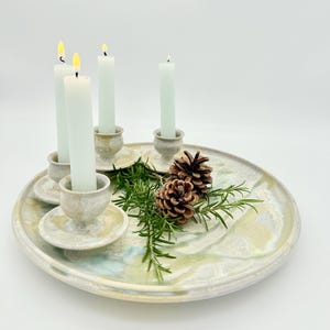 Op de afbeelding: Een groot, rond dienblad van geglazuurd keramiek met een gemarmerde afwerking in tinten crème, blauw en groen. Vier kleine, bijpassende keramische kandelaars staan op het dienblad, elk met een brandende, slanke, lichtgroene kaars.