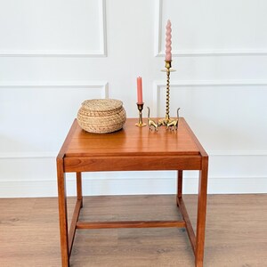 Puede incluir: Una mesa auxiliar de madera de estilo moderno de mediados de siglo con una parte superior rectangular. La mesa está adornada con una cesta tejida, dos figuras de gato de latón y dos velas rosas en candelabros de latón. La mesa mide 61 cm de largo, 46 cm de ancho y 51 cm de alto.