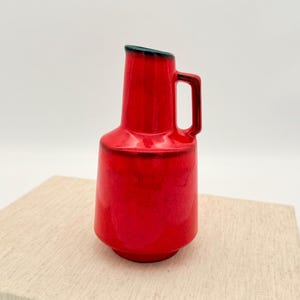 以下が含まれることがあります： 鮮やかな赤色のセラミックピッチャー。ハンドルと濃い緑色の縁が付いています。ピッチャーは円筒形の本体で、底が広がっています。ピッチャーの高さは約20cmです。