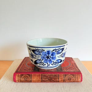 Könnte beinhalten: Eine Keramikschale mit blauem Blumenmuster. Die weiße Schale hat blaue Blumen und Blätter. Sie steht auf einem roten Buch mit goldgeprägten Mustern.