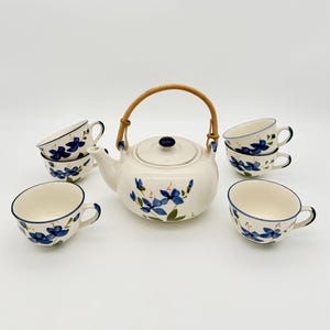 Op de afbeelding: Een keramische theeset bestaande uit een ronde theepot met een gebogen bamboe handvat en bijpassend deksel, vergezeld van vijf theekopjes. Elk stuk is gebroken wit met een handgeschilderd blauw bloemmotief, groene bladeren en dunne blauwe randen.