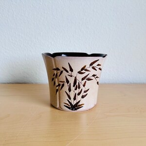 May include: A ceramic planter with a flared, scalloped rim and a light beige exterior. The body features a dark brown painted design of bamboo stalks and leaves. The interior of the planter is finished in a solid, dark brown glaze.