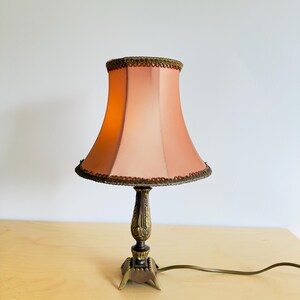 Pu&ograve; includere: Una lampada da tavolo vintage con un paralume color pesca e una base decorativa in ottone. Il paralume ha un bordo smerlato. La lampada &egrave; su una superficie di legno chiaro. Il cavo della lampada &egrave; visibile.