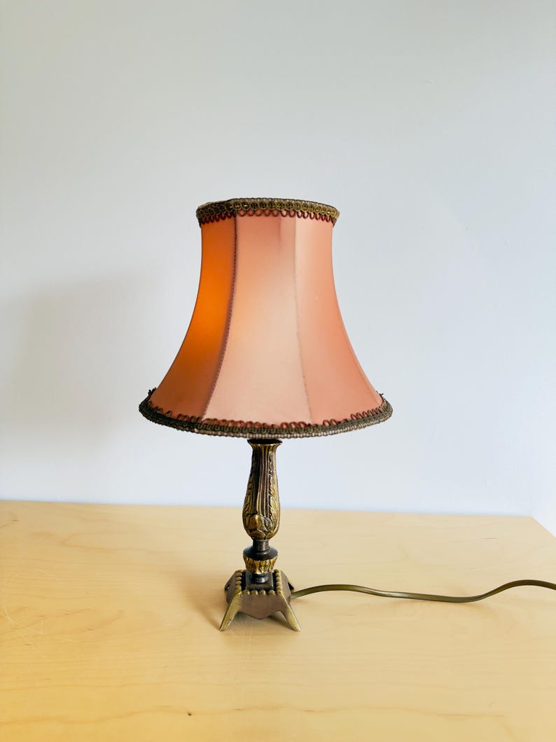 Pu&ograve; includere: Una lampada da tavolo vintage con un paralume color pesca e una base decorativa in ottone. Il paralume ha un bordo smerlato. La lampada &egrave; su una superficie di legno chiaro. Il cavo della lampada &egrave; visibile.