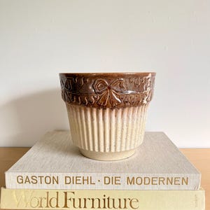 May include: A two-tone ceramic planter with a brown, embossed floral design at the top and vertical ribbed detailing on the lower half. The planter sits atop a stack of books with gold lettering.