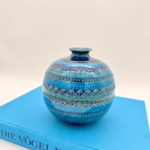 May include: A spherical, blue ceramic vase with a narrow neck and detailed geometric patterns. The vase is in shades of turquoise and dark blue, with a glossy finish. It rests on a blue book.