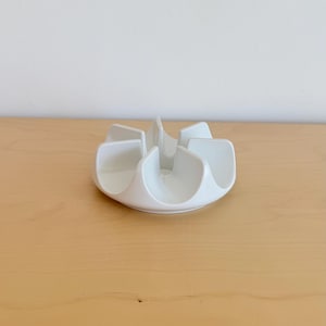 May include: A white ceramic serving dish with a unique, petal-like design. The dish has six curved sections radiating from a central point, creating a flower-like shape. The glossy finish reflects light, highlighting the smooth, clean lines of the piece.