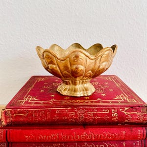 Könnte beinhalten: Eine goldfarbene Schale mit gewelltem Rand steht auf einem Stapel roter, antiker Bücher. Die Schale hat einen Sockel und eine strukturierte Oberfläche. Die Bücher haben goldene Schrift und dekorative Muster.
