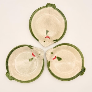 Könnte beinhalten: Drei keramische Teller in Fischform. Jeder Teller hat einen beigefarbenen Innenraum, einen grünen Rand und ein Fischgesicht-Design mit rotem Mund und grünem Auge. Die Teller sind in einem Dreiecksmuster auf weißem Hintergrund angeordnet.