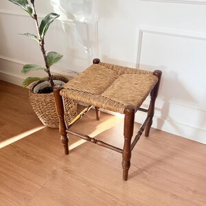Op de afbeelding: Een vierkante kruk met een donkerbruin houten frame met gedraaide poten en een gevlochten zitting van biezen. De kruk heeft vier verticale poten die verbonden zijn door horizontale dwarsbalken, wat zorgt voor een stabiele basis voor de zitting.