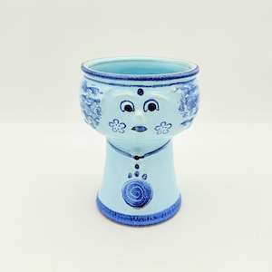 Könnte beinhalten: Ein hellblauer Keramik-Pflanztopf mit einem skurrilen Gesichtsdesign. Der Pflanztopf hat eine runde Oberseite mit blauem Rand, ein Gesicht mit Augen, Nase und Mund sowie einen stilisierten Körper. Der Pflanztopf ist etwa 15 cm hoch.