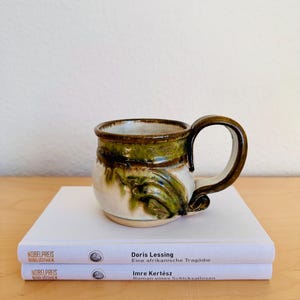 Könnte beinhalten: Ein Set aus drei Keramikbechern mit gesprenkelter, matter Oberfläche. Die Becher sind in den Farben Creme, Hellgrau und Dunkelgrau gehalten. Jeder Becher hat ein schlichtes, minimalistisches Design und einen abgerundeten Henkel.