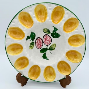 Può includere: Un piatto in ceramica bianca con una trama intrecciata, bordo verde e un disegno centrale di frutta e foglie. Il piatto è decorato con disegni gialli a forma di uovo sul bordo. Il piatto è esposto su un supporto di legno.