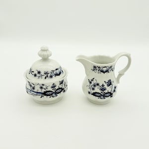 Peut inclure: Un sucrier en céramique blanche avec couvercle et un pot à crème. Les deux pièces sont décorées d'un motif floral bleu. Le sucrier est rond et le pot à crème a une anse et un bec verseur.