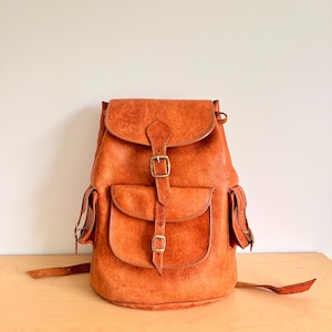 Puede incluir: Una mochila de cuero marrón con dos bolsillos delanteros y un cierre de solapa asegurado con hebillas. La mochila tiene correas ajustables y un asa superior. El cuero tiene una apariencia texturizada.